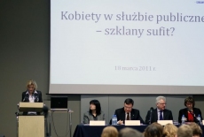 Pani stoi przy mównicy i przemawia, obok siedzą Paneliści przy stole 