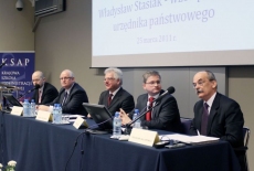 Paneliści siedzą przy stole prezydialnym, pan mówi do mikrofonu