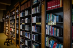 Regał w bibliotece na którym stoją książki. Obok stoi stołek.