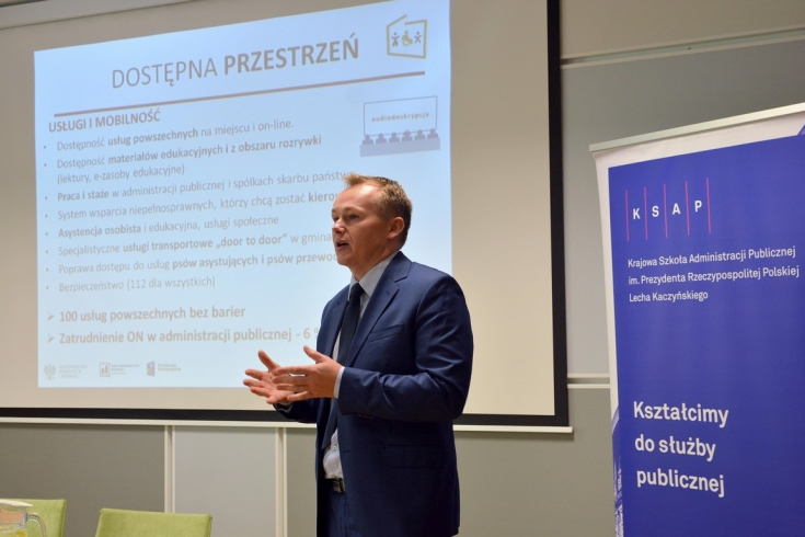 Prelegent podczas wystąpienia, w tle na ekranie wyświetlona prezentacja "Dostępna przestrzeń"